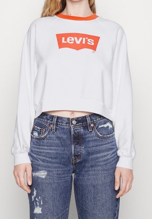 Person trägt ein weißes langärmliges Levi's Cropped Sweatshirt mit orangefarbenem Logo und Kragen, kombiniert mit blauen, abgenutzten Jeans.