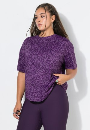Joven mujer con cabello largo que lleva una camisa morada con estampado de leopardo y pantalones morados oscuros, mirando hacia un lado con una expresión neutral.
