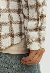 Camisa de franela con un patrón de cuadros en blanco y marrón, que presenta un puño abotonado. Lleva puesta sobre una camiseta beige, combinada con unos vaqueros azules.