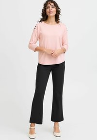 Haut rose clair à manches trois quarts avec un tissu texturé et des accents de boutons noirs sur les épaules ; associé à un pantalon noir évasé et des chaussures beiges.