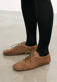 Rjave nubuk čevlje z vezalkami in ravnim podplatom, ki imajo šivane detajle in okroglo konico, nosijo z črnimi treginkami. Gladka tekstura, sproščen dizajn.