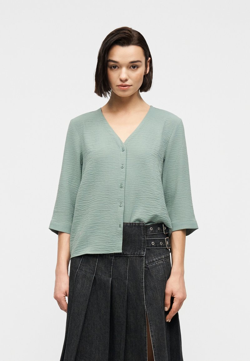 Γυναίκα με κοντά σκούρα μαλλιά, φορώντας μπλούζα χρώματος φασκόμηλου με υφή, κουμπωτή, με μανίκια τριών τετάρτων και ψηλόμεση πλισέ φούστα από σκούρο τζιν.