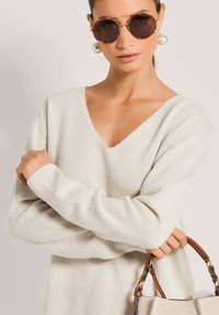 Hellgrauer, langärmeliger Strickpullover mit V-Ausschnitt und gerippten Bündchen. Das Modell hält eine Handtasche mit strukturiertem Obermaterial und Lederakzenten.