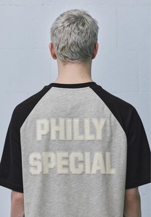 Osoba z krótkimi siwymi włosami, ubrana w czarno-szarą koszulkę z widocznym napisem "PHILLY SPECIAL" na plecach.