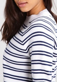 Donna che indossa un maglione bianco con strisce orizzontali blu navy equidistanti, mostrata in profilo laterale dalle spalle alle labbra.