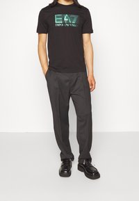 Svart t-shirt med grön grafisk logotyp, matchad med skräddarsydda grå byxor och svarta skor. Slät tyg och figursydd design.