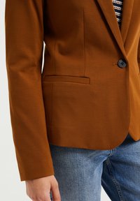 Brun kavaj i mjukt tyg med en enda svart knapp, slagkrage och sidoficka. Matchad med blå denimjeans.