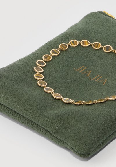 Collar en tono dorado con cuentas de ojo de tigre en forma ovalada, descansando sobre una bolsa de ante verde con el texto en relieve "JIAJIA". Textura suave, diseño compacto.