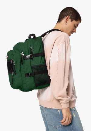 Sac à dos en nylon vert avec plusieurs compartiments, poche en mesh sur le côté, sangles d'épaule réglables et détails de fermeture à boucle noire.