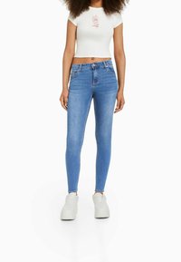 Kvinna klädd i högmidjade ljusblå skinny jeans, vit crop top med rosdesign och vita platåskor, står framför vit bakgrund.