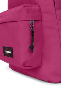 Rosa tygryggsäck med en sidoficka, som har en svart dragkedja och en svart Eastpak-logotyp. Slät textur och kompakt form.