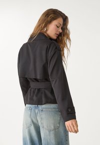 Svart bältejacka med en kortad design och strukturerade axlar, med en bakre flik och knäppning vid ärmsluten. Buren med ljusblå denim.