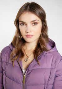 Jachetă puf purple cu fermoar pe partea din față și glugă, având o capitonare orizontală. Hardware-ul auriu și o colier cu pandantiv floral sunt, de asemenea, vizibile.