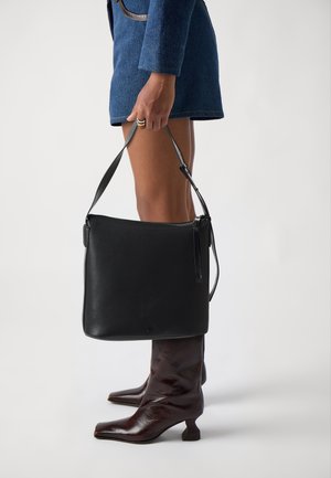 Persona con gonna di jeans e stivali marroni con tacco che tiene una borsa a tracolla nera in pelle per la tracolla di lato.
