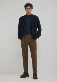Maglione zip-up a coste blu scuro sopra una maglietta blu navy, abbinato a pantaloni marroni e stivali in suede marroni. Posizione autonoma davanti a uno sfondo neutro.