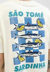Cremfarbendes T-Shirt mit einem Motiv von vier Fischen mit Zitronenscheiben auf einem blau-weiß karierten Hintergrund. Der Text lautet "SÃO TOMÉ SARDINHA."