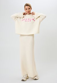 Lyhyt, kermaisen valkoinen neulepaita, jossa pinkillä teksti "Good Karma Club"; yhdistetty pitkään, vartaloa myötäilevään hameeseen. Pehmeä neulos. Rentoon tyyliin sopivat jalkineet.