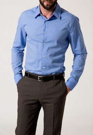 Mężczyzna ubrany w jasnoniebieską koszulę na guziki wpuszczoną w ciemnoszare spodnie garniturowe z czarnym paskiem, stojący z rękami w kieszeniach.