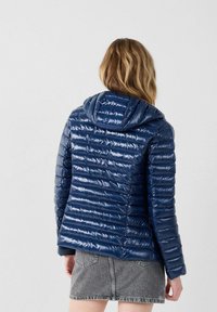 Giubbotto imbottito blu navy con finitura lucida, caratterizzato da sezioni cucite orizzontalmente e un cappuccio. Stoffa testurizzata e design aderente.