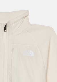 Κρεμ φλις μπουφάν με ψηλό γιακά και φερμουάρ μπροστά, με λευκό λογότυπο "The North Face" στο στήθος.