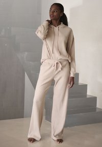 Bežani rebrasti loungewear set s kapuljačom i hlačama s vezicom. Mekana tekstura, labav kroj i vertikalni uzorak.