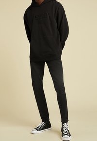 Persona che indossa un hoodie nero con logo in rilievo, jeans neri aderenti e sneakers alte nere con lacci bianchi, in piedi su uno sfondo beige.