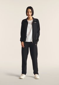 Giacca nera con zip e dettagli dorati, t-shirt bianca con motivo a taglio, e pantaloni jogger neri. Il modello indossa sneakers bianche.