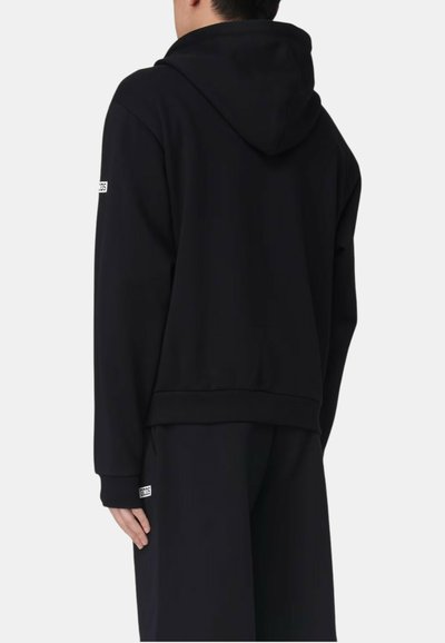 Osoba ubrana w czarną bluzę z kapturem i czarne spodnie, pokazująca widok od tyłu, z małymi białymi naszywkami z logo na rękawie i spodniach.