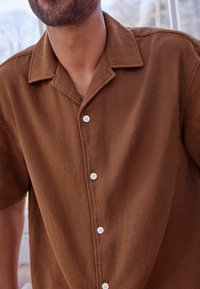 Brązowa koszula z krótkim rękawem z teksturowanej tkaniny, z tradycyjnym kołnierzem i białymi guzikami z przodu. Delikatnie widoczne detale przeszyć.