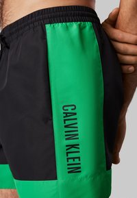 Szorty kąpielowe w kolorze czarnym i jaskrawej zieleni, wykonane z gładkiego materiału. Posiadają elastyczny pas i nadrukowaną nazwę marki w kolorze czarnym wzdłuż boku.