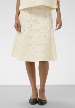 Femme portant une jupe midi crème à œillets et des ballerines pointues noires, debout avec les mains détendues sur les côtés sur un fond uni.