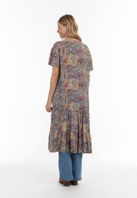 Maxime puku yenye mchoro wa paisley kwa buluu, shani na manjano, mikono mifupi, muundo unaotembea, na hem iliyoteleza, ikiunganishwa na suruali za denim.