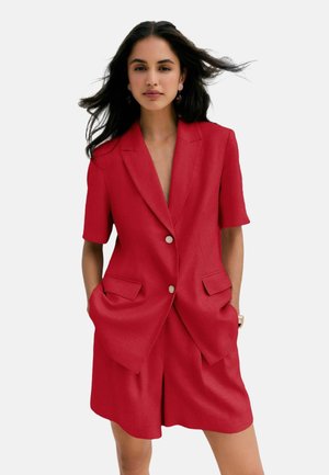 Žena s dlhými tmavými vlasmi nosí červený blazer s krátkym rukávom s gombíkmi a zodpovedajúce na mieru šité šortky, stojí s rukami v vreckách.