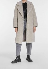 Lysegrå, blød shearling-frakke med stor krave og knaplukning. Båret over sort skjorte og grå jeans, parret med sorte ankelstøvler.
