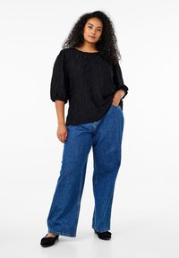 Black textured blouse with wide sleeves, paired with loose blue denim pants. Footwear is black flats. Neutral background.