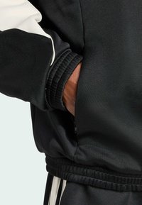 Jaqueta atlética preta com detalhes em branco, um bolso com zíper na lateral, punhos elásticos e textura que indica material sintético.