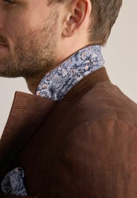 Braune Jacke mit einem blauen, floral gemusterten Kragen. Der Kragen weist komplizierte Muster in Blau- und Orangetönen auf, die der Gesamtgestaltung Textur verleihen.