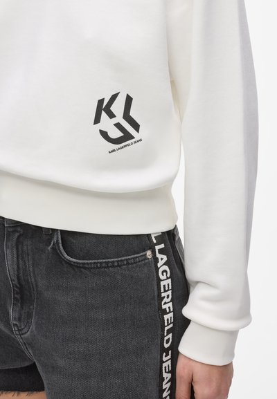 Бял суичър с черно лого "KLJ Karl Lagerfeld Jeans" над подгъва, комбиниран с черни дънкови къси панталони с лента с лого отстрани.