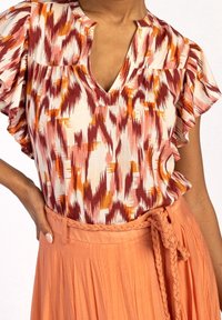 Blusa stampata con maniche corte a volant in tonalità di rosso, arancione e crema. Abbinata a una gonna arancione pieghettata e leggera.