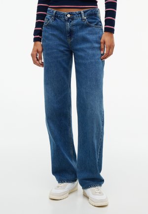 Personne portant un jean bleu à jambes larges avec bouton et fermeture éclair, associé à des baskets blanches et un top court à manches longues rayé bleu marine.