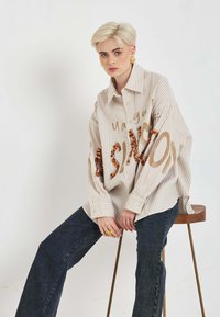 Pruhovaná béžová oversized košeľa s hnedými flitrovanými nápismi. Má gombíkový golier a dlhé rukávy. Nosí sa s tmavými džínsami.