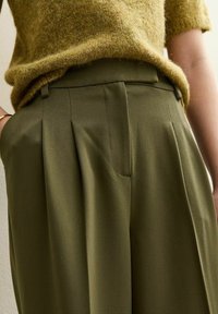 Pantalones plisados de color verde oscuro con una textura suave, que presentan una cintura alta y un ajuste entallado, combinados con una blusa de punto ligera en color mostaza.