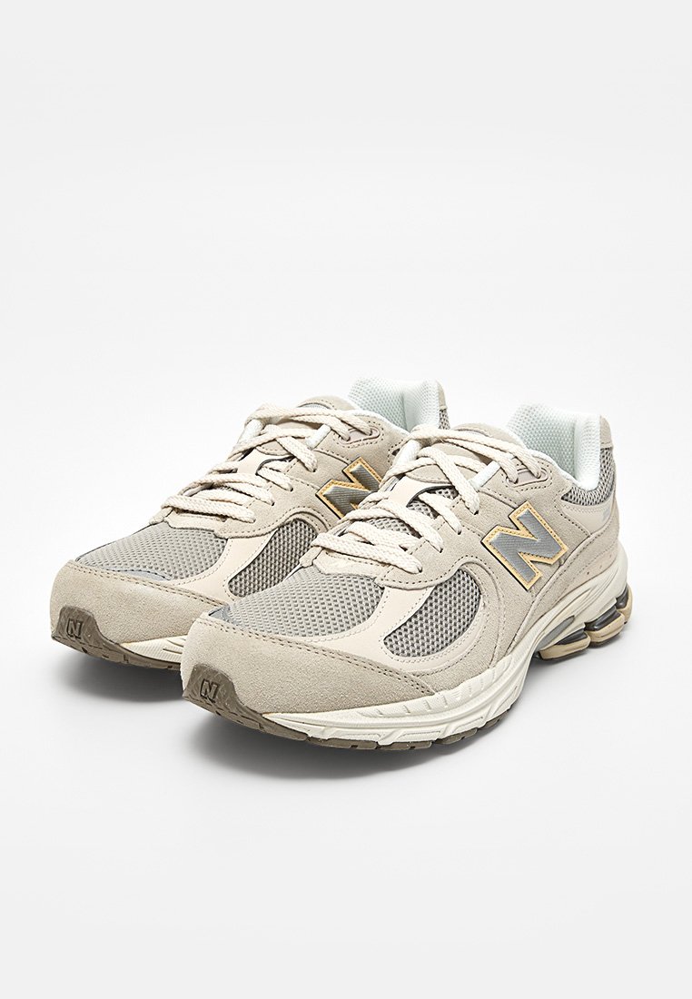 Coppia di sneakers New Balance beige e grigie con pannelli in mesh e pelle scamosciata, chiusura con lacci e suole imbottite su sfondo bianco.
