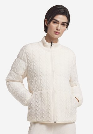 Femme portant un pull crème en maille torsadée avec fermeture éclair et poches, posant les mains dans les poches sur un fond uni.