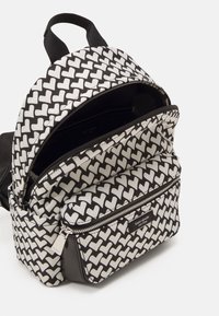 Svart och vit ryggsäck med ett upprepande hjärtmönster, slät tyg, dubbla blixtlåsfack och en logotyp på framsidan.