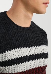 Pull en maille noir, blanc et bordeaux. Présente un motif texturé avec des rayures horizontales. Col rond, coupe ajustée.