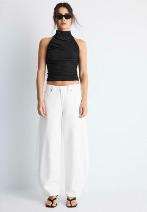 Jonge vrouw draagt een zwarte mouwloze gerimpelde top, witte brede broekspijpen, zwarte sandalen en zwarte zonnebril, staand tegen een witte achtergrond.