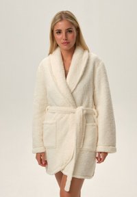 Kremowy, pluszowy szlafrok z kołnierzem szalowym, paskiem i dwoma przednimi kieszeniami. Miękka tekstura i średnia długość. Brak wzorów i widocznych elementów metalowych.