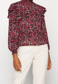 Blommig blus med volangärmar; svart bas med röda och rosa blommor; hög krage; lätt tyg; långa ärmar med elastiska manschetter.