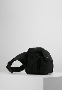 Svart midjeväska med flera fickor och snäpplock, placerad på en vit pedestal mot en enkel grå bakgrund.